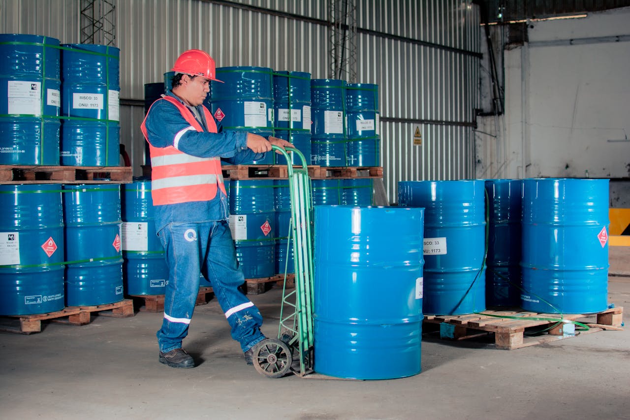 get-in-touch Industrial worker with a hand truck moving blue barrels in a warehouse setting.